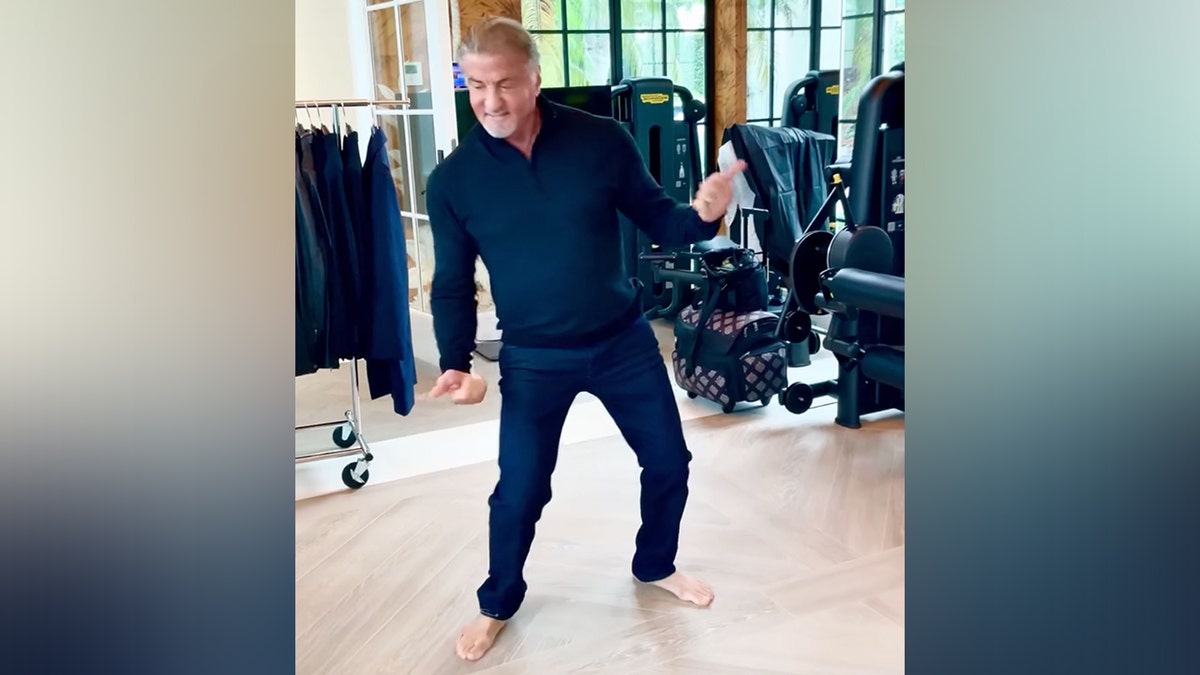 Sylvester Stallone dengan celana dan kemeja biru bersandar ke samping saat dia menari