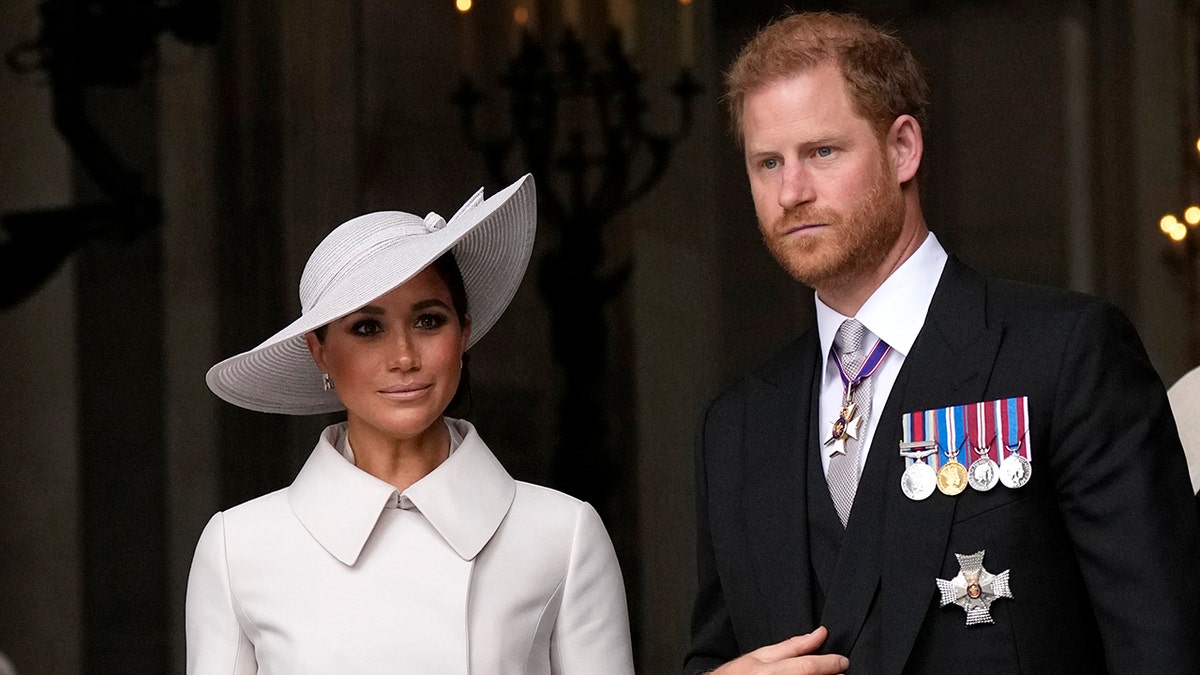 Pangeran Harry dalam setelan hitam dengan medali dan topi serasi dalam jas putih di samping Meghan Markle.