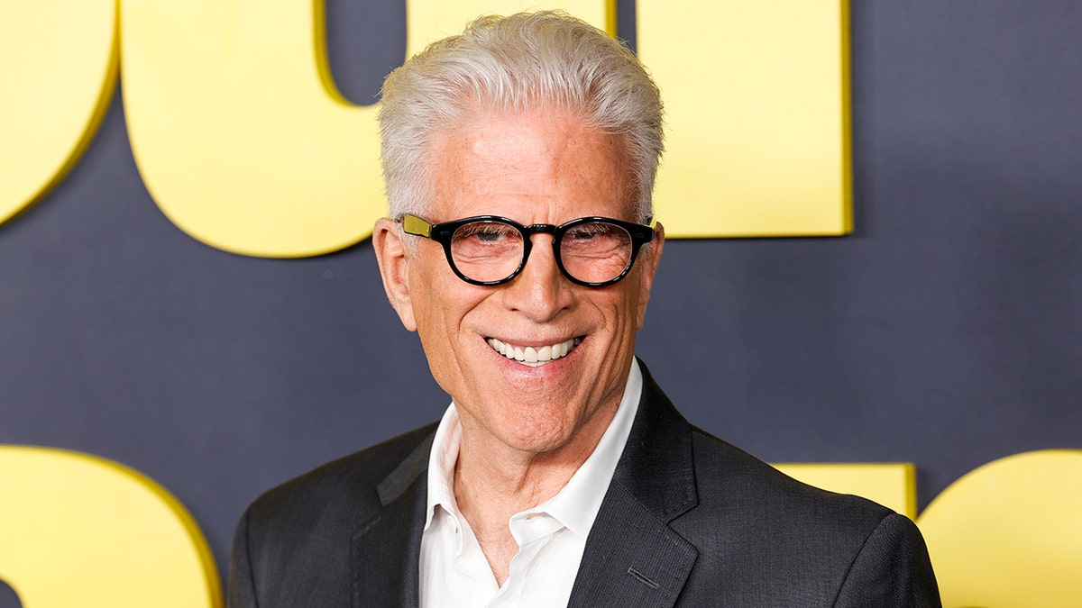 Ted Danson dengan kemeja putih dan blazer hitam dengan kacamata bulat hitam tersenyum di atas karpet