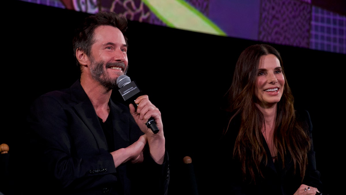 Keanu Reeves tersenyum di atas panggung sambil memegang mikrofon di wajahnya, duduk di sebelah Sandra Bullock yang tersenyum, keduanya berkulit hitam.